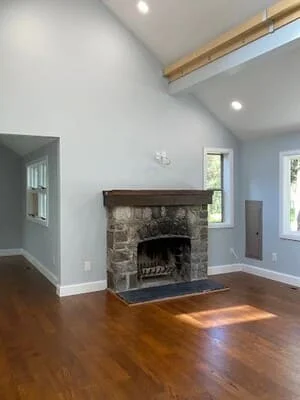 Vaulted living room with stacked stone fireplace, dark wood mantel, exposed ceiling beams, and refinished hardwood floors