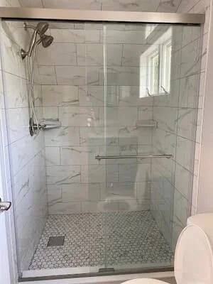 Walk-in shower with white marble-look wall tile, hexagonal mosaic floor tile, frameless glass sliding door, and chrome grab bar