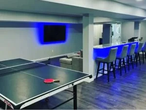 Finished basement game room with ping pong table, wall-mounted TV, blue LED-lit bar counter, and upholstered bar stools