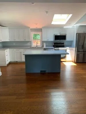 Renovated kitchen with white shaker cabinets, blue painted island, stainless appliances, and hardwood floors under a skylight