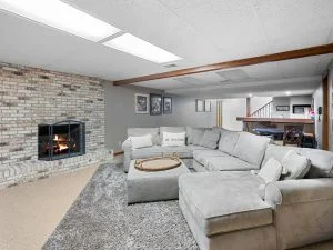 Finished basement with whitewashed stone fireplace, gray sectional sofa, shag rug, and exposed wood ceiling beam