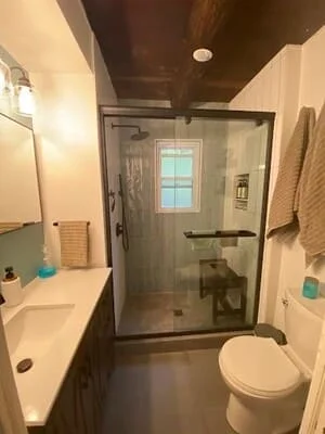 Bathroom with glass sliding shower enclosure, dark wood bench, tiled walls, and white vanity with undermount sink