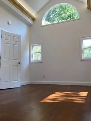 Empty room with vaulted ceiling, exposed wood beam, arched window, dark hardwood floor, and white six-panel door